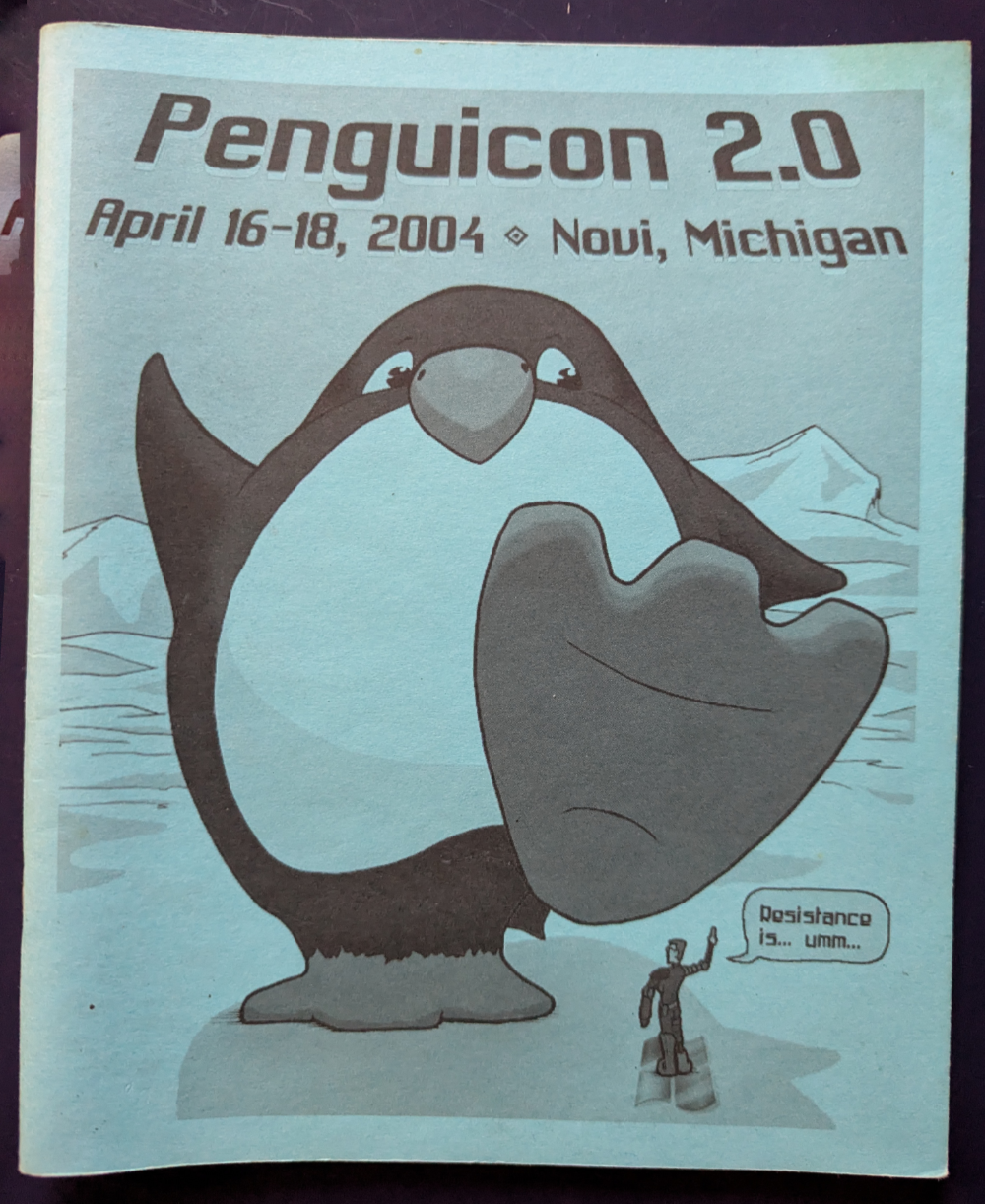 A Penguicon 2004 schedule book cover by Howard Tayler, depicting a giant Linux penguin about to step on Microsoft's Bill Gates who is dressed as the Borg from Star Trek, who is saying "Resistance is... umm..."
